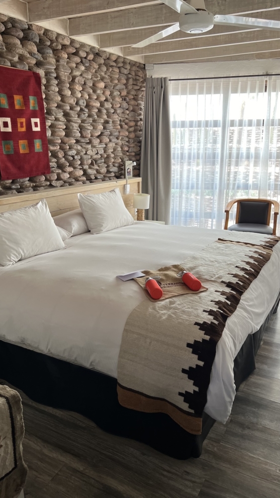 Photo of a double bed against a wall. The bed has white sheets and bed covers. The wall behind the bed is made of stones. On top of the bed is a sack with two water bottles laying down on top of it.