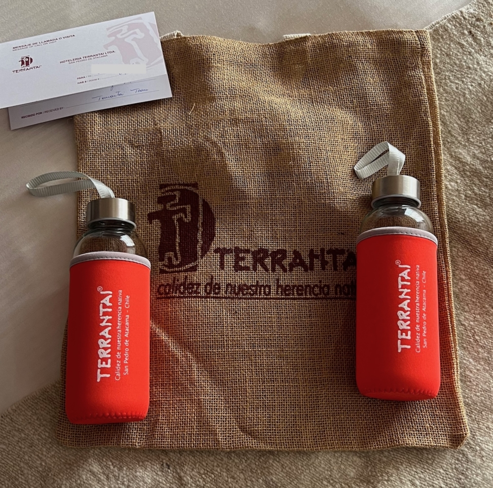 A burlap bag is on a bed. On top of the bag are two water bottles laying down. The water bottles are made of glass, and have a cover over them that have the name of the hotel, Terrantai.