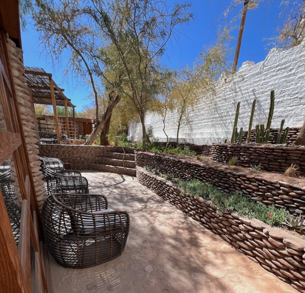 Photo of an outdoor area at Terrantai Lodge. There are a few chairs on the left side of the picture. Opposite the chairs is a terraced garden that goes from the bottom right corner to the center of the frame. Beyond that, there are stairs and a tree in the background. The walls of the terraces and stairs are made of stone that are laid like bricks.