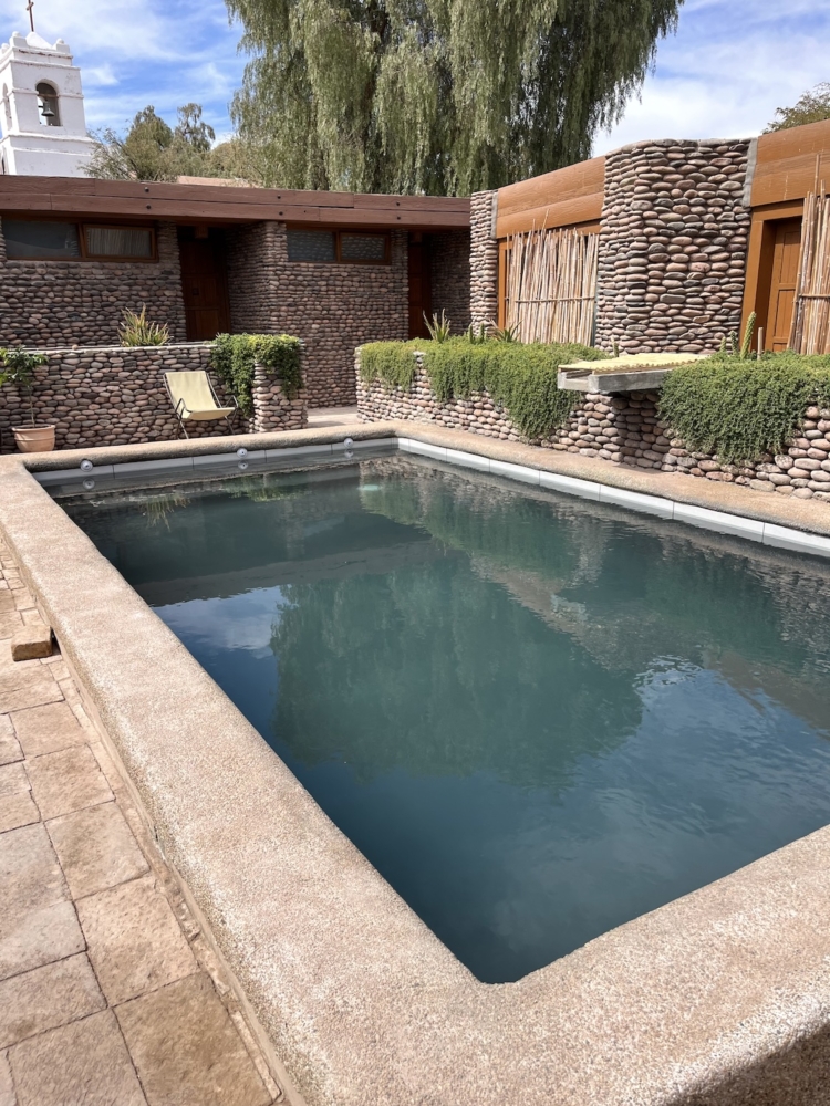 The main subject in this photo is a rectangular pool. In the background is the hotel which the walls are made of stone that are laid in a brick-like pattern. There is also a chair next to the pool. 